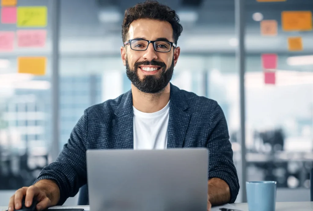 A bearded man wearing glasses sits at a desk with a laptop, symbolizing digital marketing growth services through the DCP Program